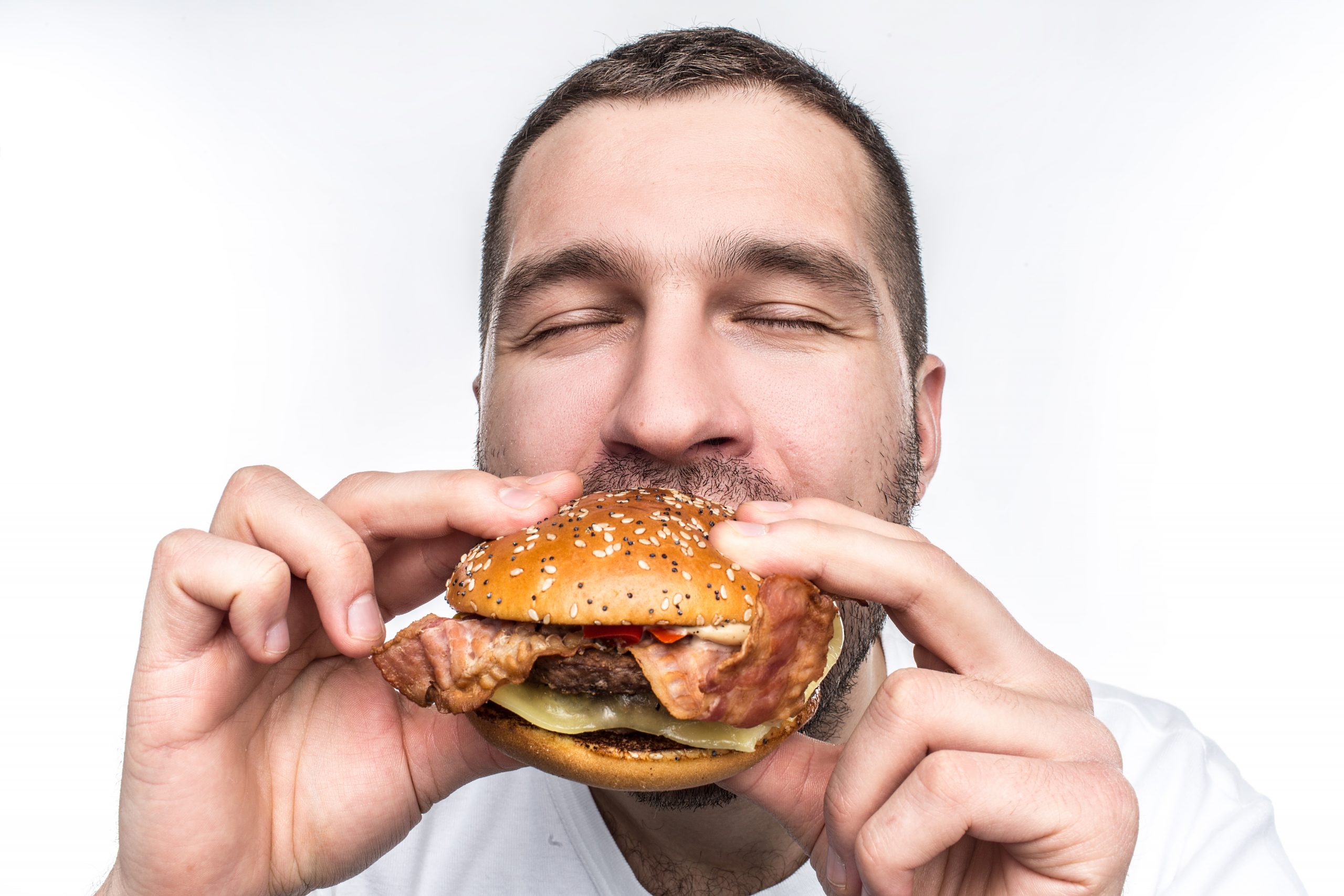 Weird and bizarre man is eating fat and juicy hamburger. It is not a healthy food but the guy likes it very much. His face is very emotional. Isolated on white background.