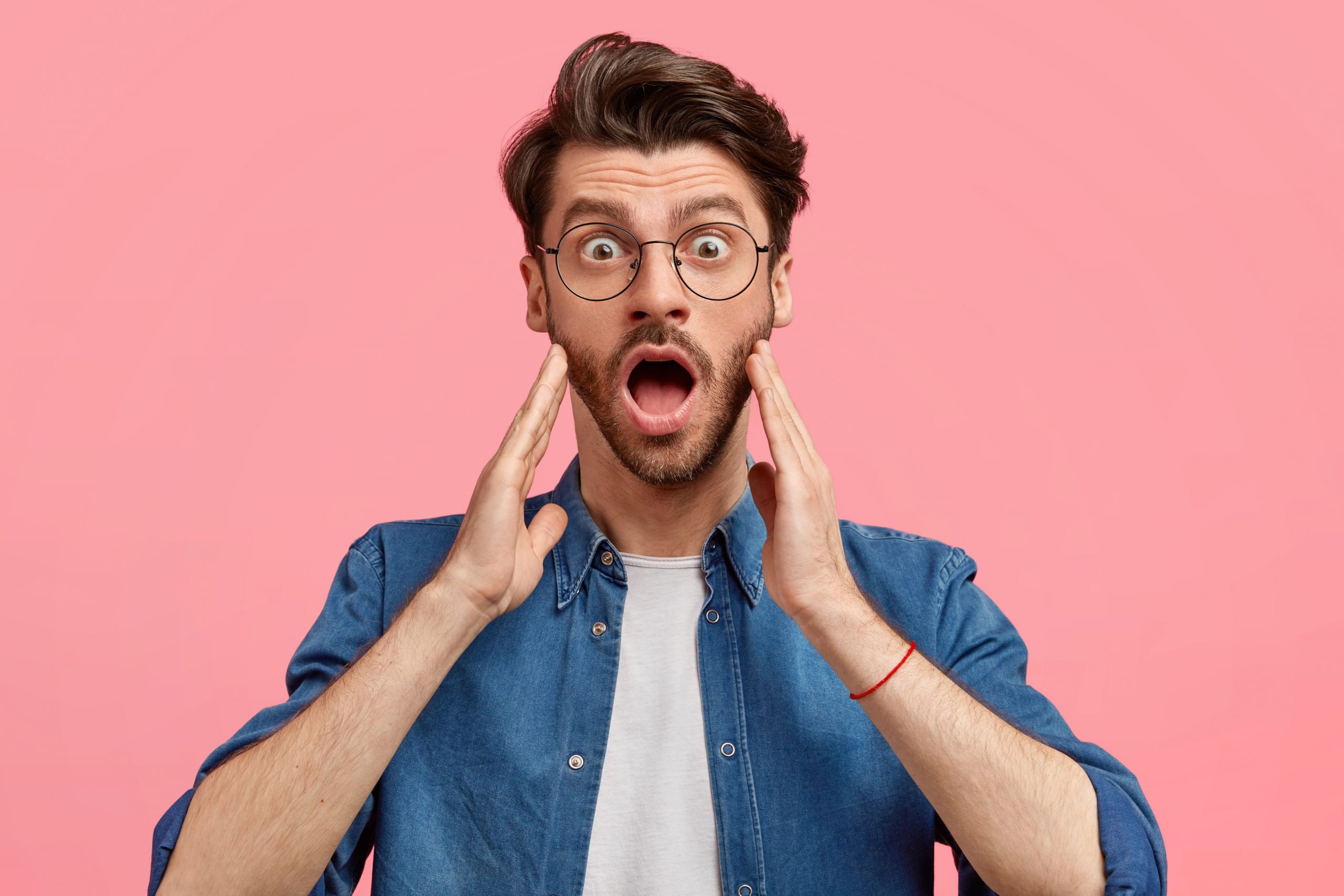 people-unexpectedness-concept-shocked-young-male-with-stubble-opens-mouth-widely-keeps-hands-cheeks-notices-something-unbelievable-wears-round-spectacles-denim-shirt-stands-indoor